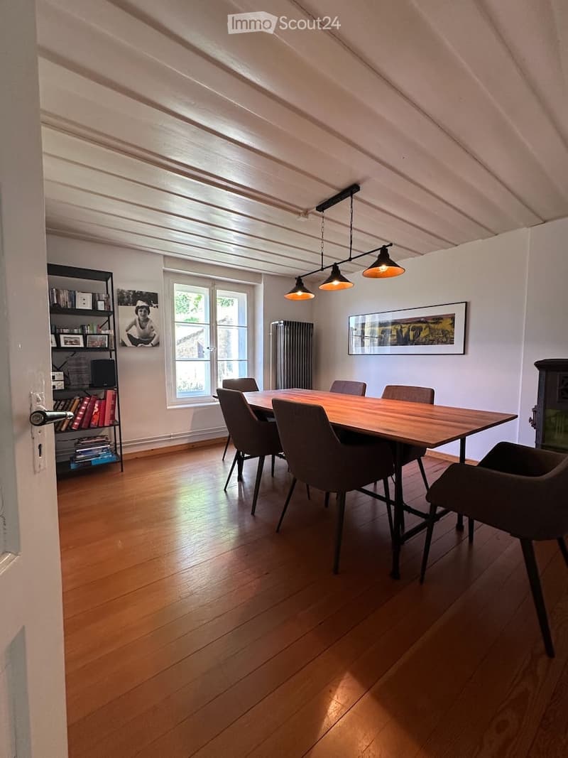 Furnished Room. Bright dining area with wooden floor, large window, modern dining table with six chairs, bookshelf, radiator, and artwork on the wall.