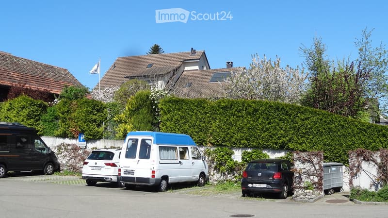 Building. Residential building with a driveway and several parked vehicles, including a van, a car, and a van, surrounded by greenery and a hedge.