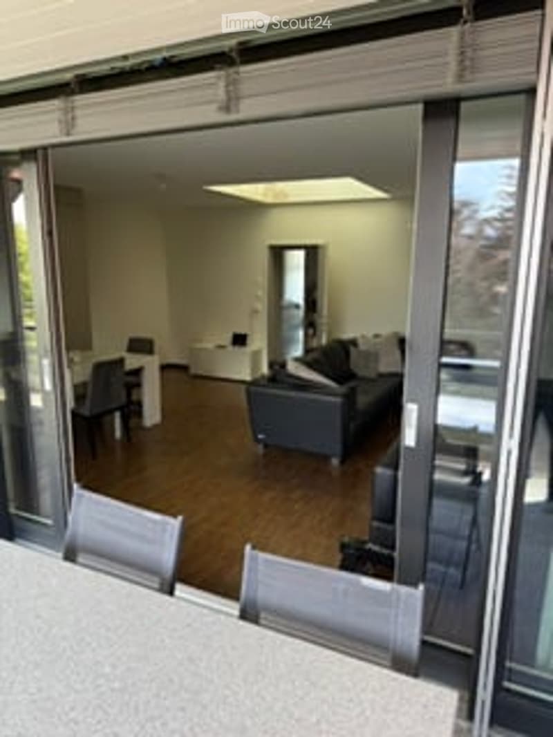 Furnished Room. Bright, modern living space with large glass sliding doors leading to a balcony, wooden flooring, a grey sofa, dining chairs, and a small table.