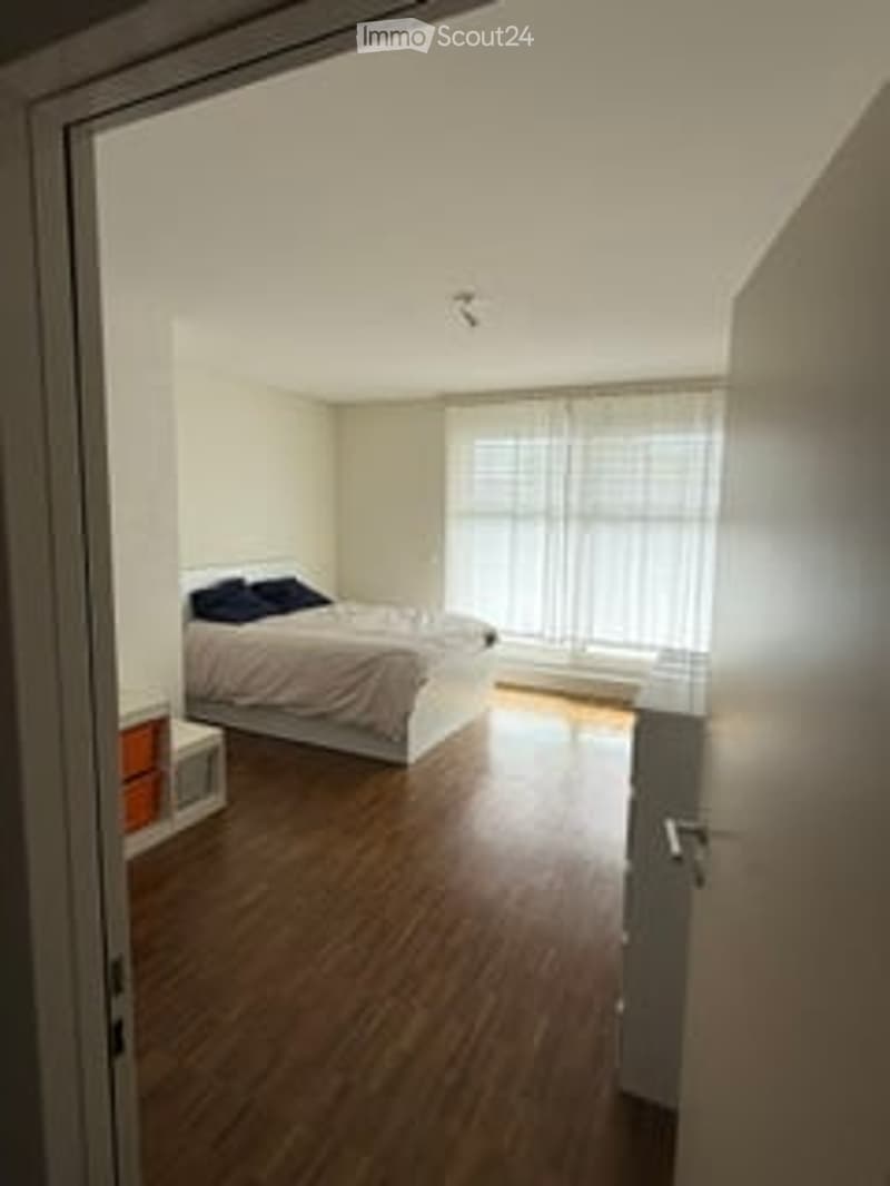 Bedroom. Bright bedroom with wooden flooring, large window with white curtains, bed with dark blue pillows, nightstand, and lamp.