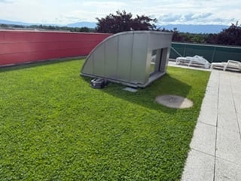 Garden / Outdoor Area. A small, modern outdoor structure with a curved roof sits on a grassy area next to a paved walkway. The surrounding area includes a red wall and distant mountain views.