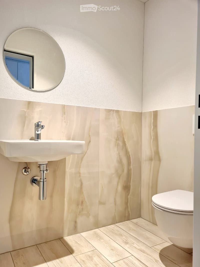 Bathroom. Modern bathroom with a white sink, chrome faucet, toilet, and a large round mirror on the wall. Light-colored marble tiles and wooden flooring.