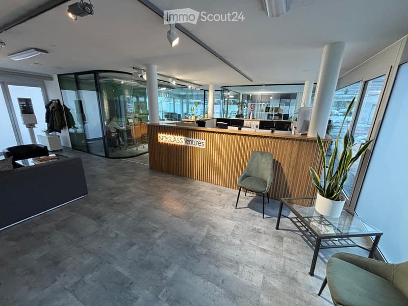 Modern office lobby with a reception desk, seating area, and large windows. Features include a sofa, chairs, a small table with a plant, and a coat rack with jackets.