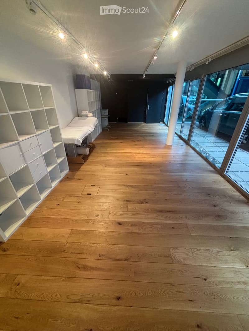 Spacious room with wooden flooring, large windows, white shelving unit, and a treatment table.