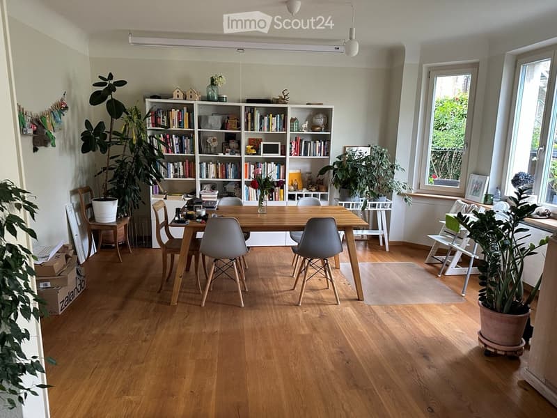 Bright, airy room with wooden flooring, large window, and a dining table surrounded by chairs. Shelves filled with books and decorative items, along with several potted plants, create a cozy and inviting atmosphere.