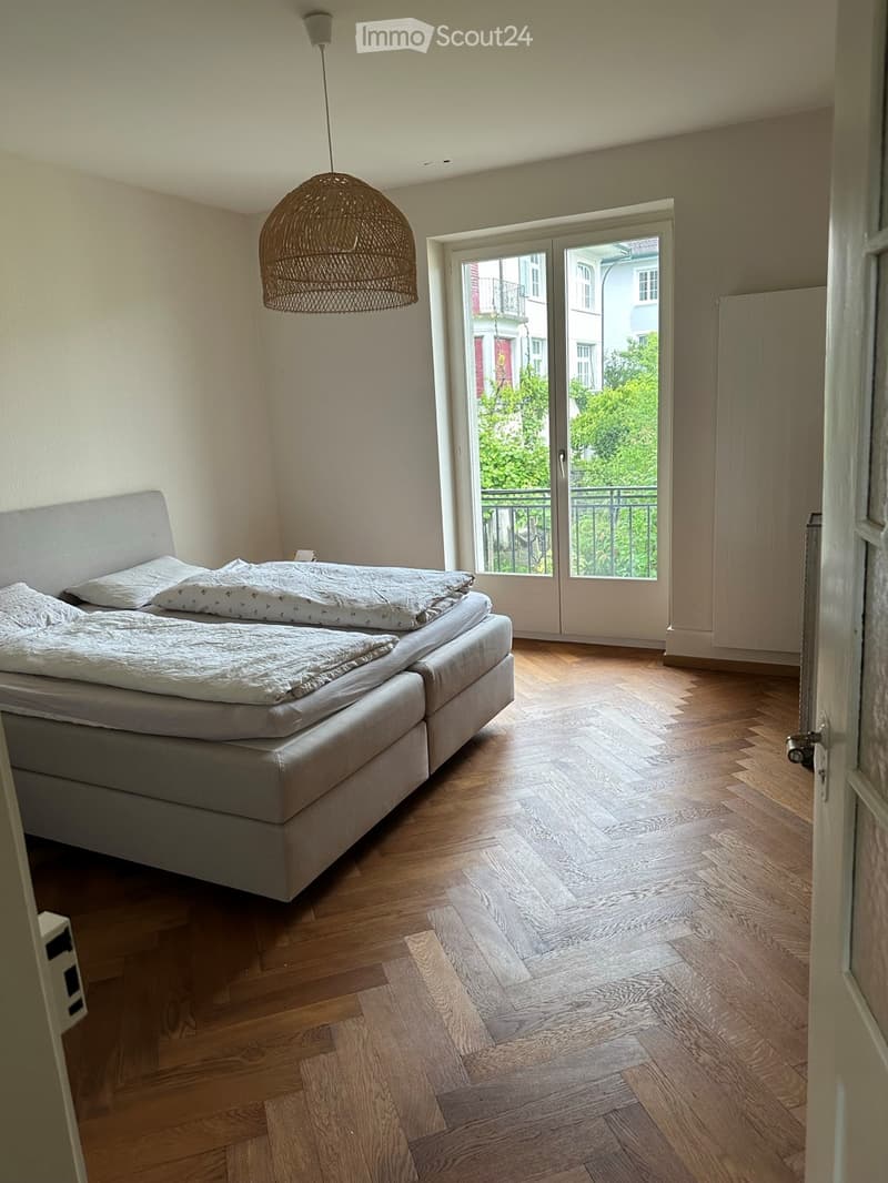 Bright bedroom with wooden flooring, a modern bed with white bedding, and a large window opening to a balcony. A stylish wicker pendant light hangs from the ceiling.