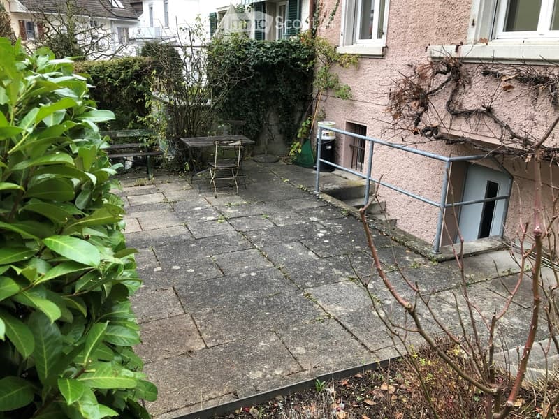 Small concrete patio area with a metal railing, a small table and chairs, and a green bush in the foreground.