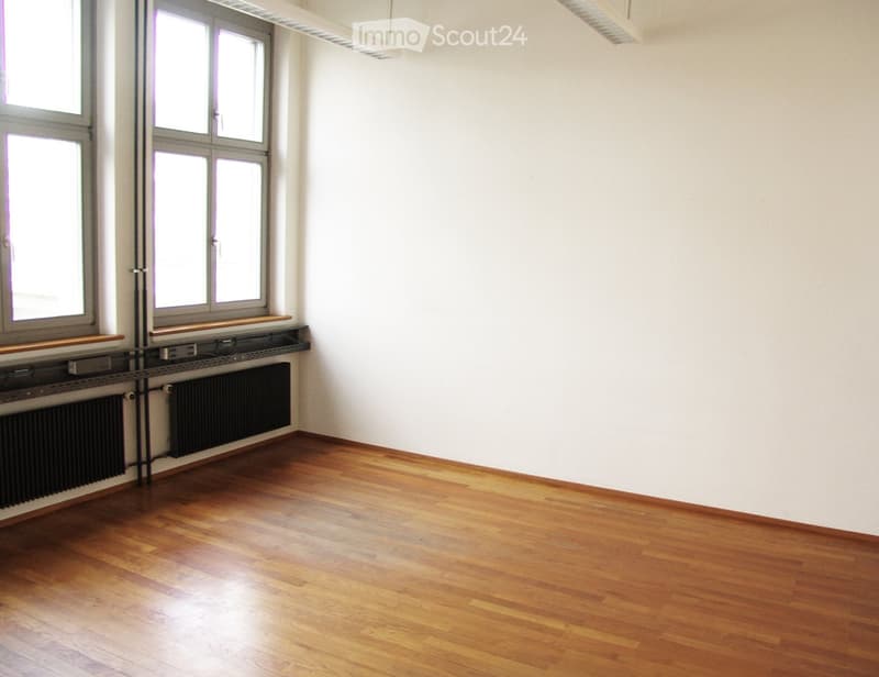 Empty room with wooden flooring, white walls, and large windows. Radiators are visible along the wall below the windows.