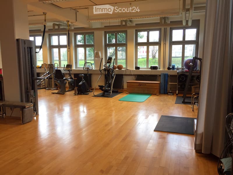Well-lit fitness room with wooden flooring, large windows, and various exercise equipment including bikes, a treadmill, weights, and yoga mats.