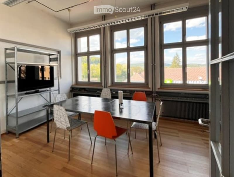 Bright room with large windows, wooden floor, a table surrounded by chairs, and a TV screen on a metal stand.