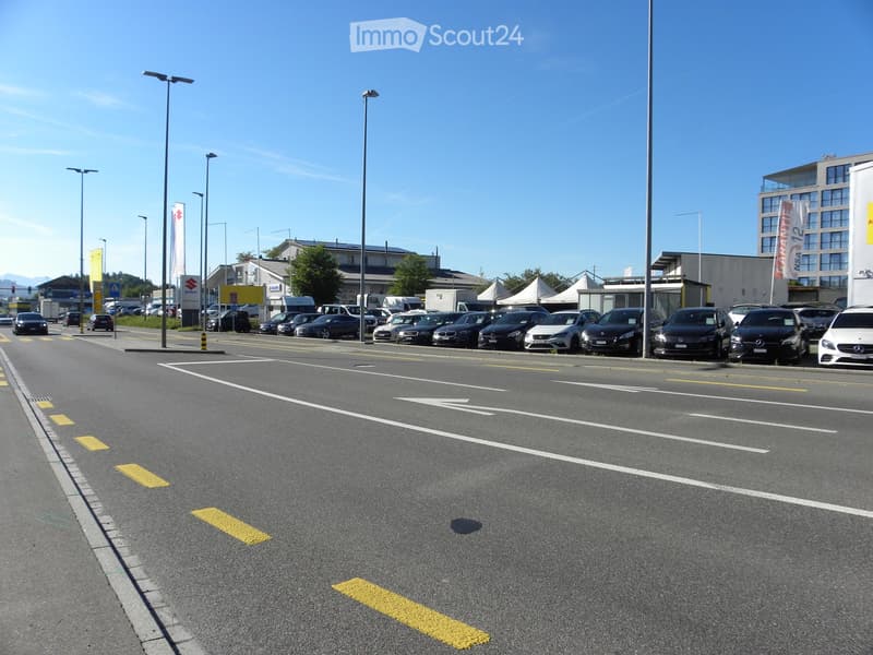 Car dealership lot with multiple parked cars, banners, and surrounding buildings.