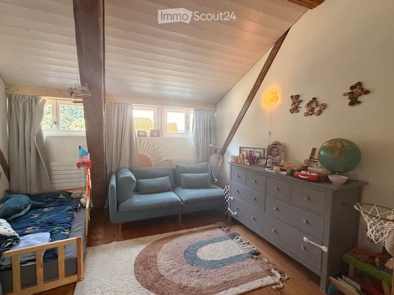 Cozy attic bedroom with slanted wooden ceiling, large windows, a blue sofa, a wooden bed, a dresser with various items, and a globe on top.