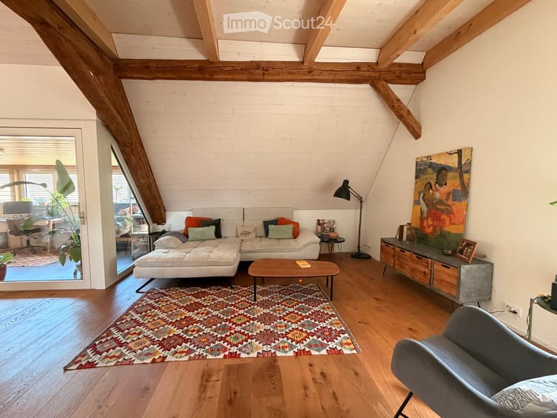 Modern living room with exposed wooden beams, white walls, wooden flooring, and large windows. Features a white sectional sofa with colorful cushions, a wooden coffee table, a side table, a floor lamp, and a grey armchair.