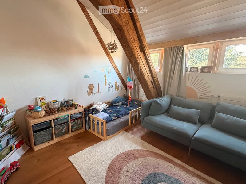 Cozy attic bedroom with wooden beams, a blue sofa, a small bed, a storage unit, and various toys and decorations.