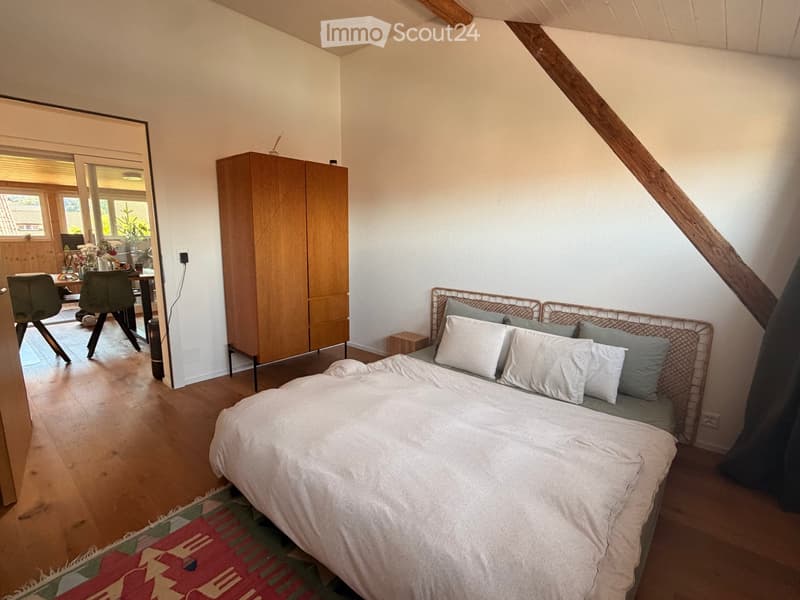 Cozy attic bedroom with wooden floors, slanted ceilings, a large bed, a small sofa, a dining area with chairs and a table, and a wooden cabinet.