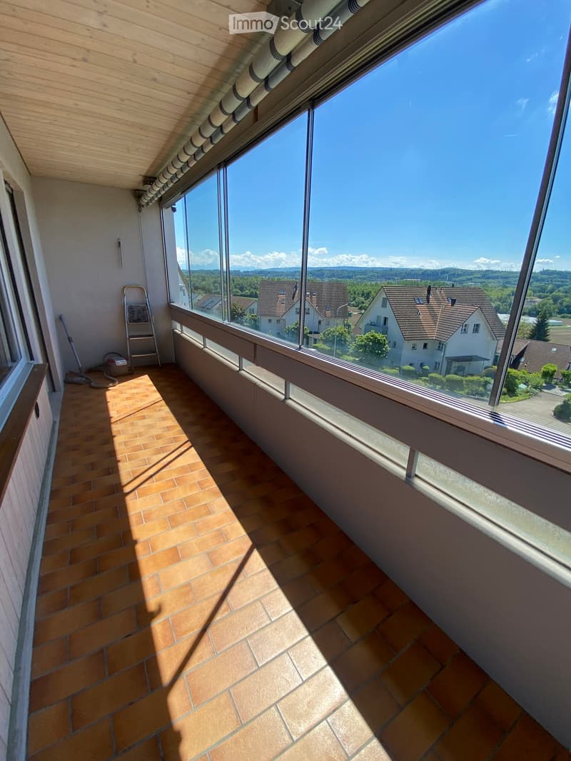 Balcony with large windows, tiled floor, and a view of surrounding houses and landscape.