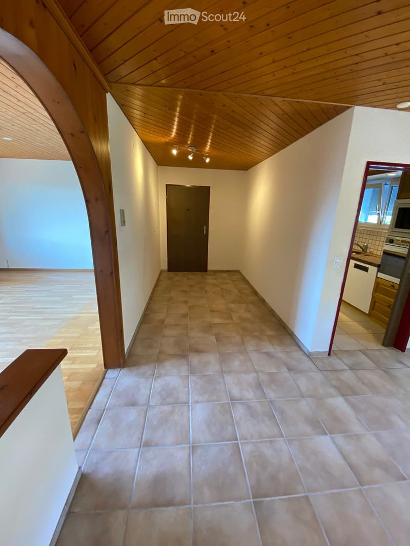 Empty hallway with tiled floor, wooden ceiling, and a closed door at the end. Natural light from the left side and a glimpse of a kitchen area on the right.