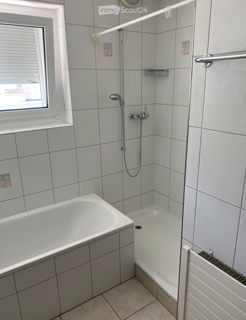 Modern bathroom with a bathtub, shower area, tiled walls, and a window with blinds.