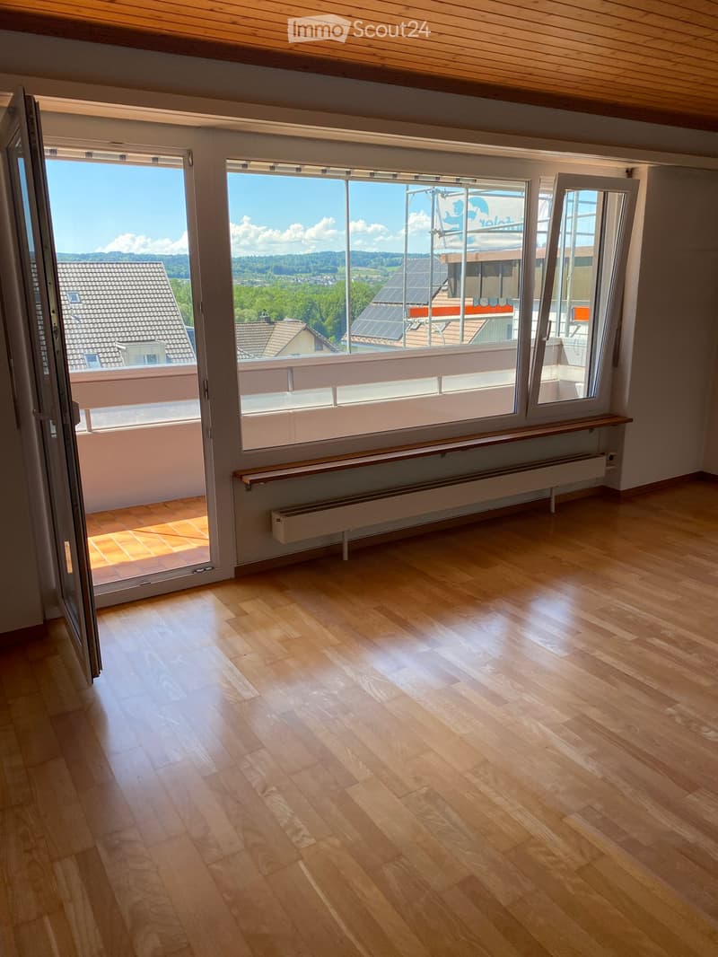 Empty room with wooden flooring, large windows, and a balcony area visible through the open sliding doors.