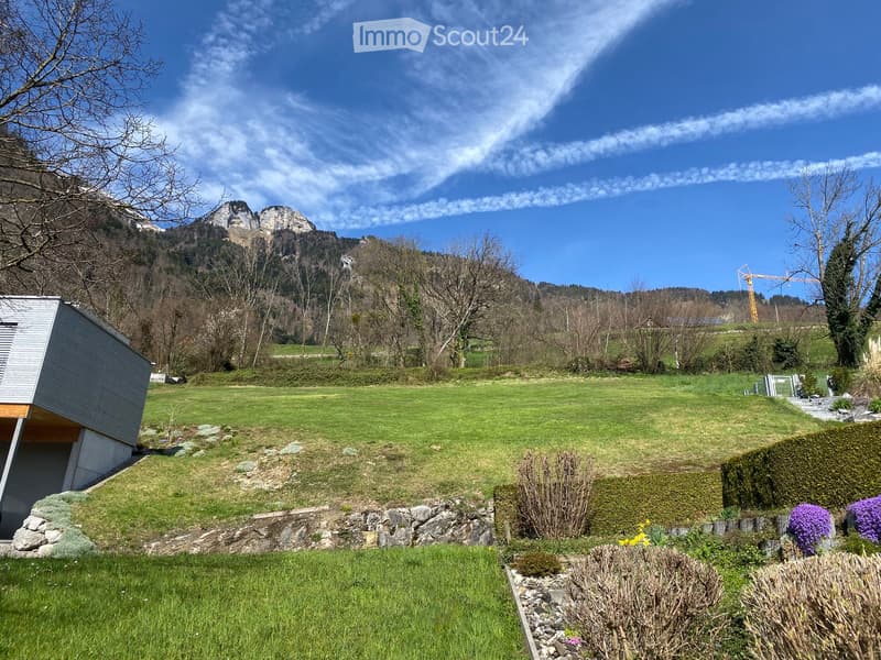 A well-maintained garden with a modern house on the left, surrounded by a grassy lawn, shrubs, and a stone wall. The background features a mountain range under a clear blue sky.