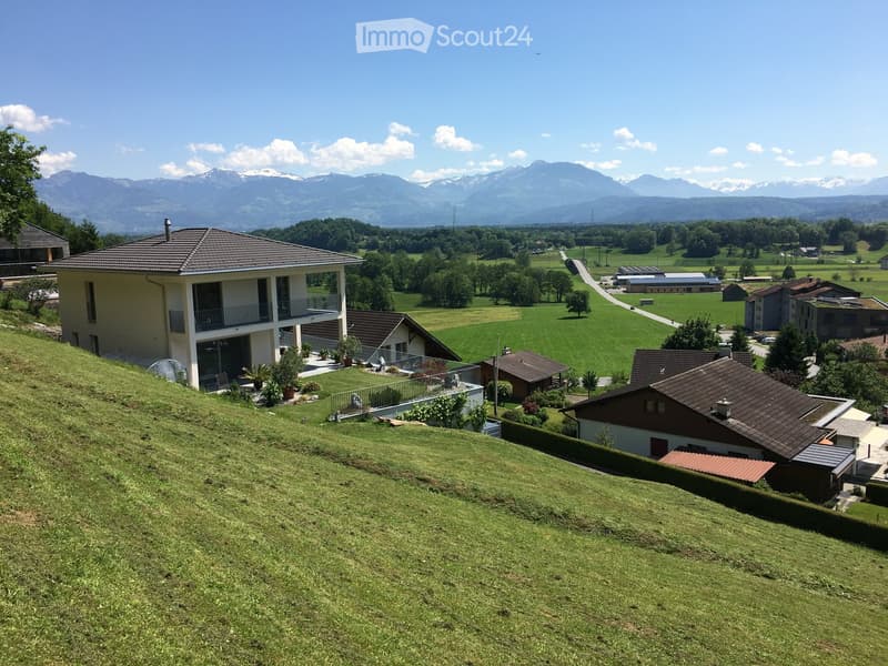 A two-story house with a balcony and a garage, situated on a grassy hillside with a scenic mountain view in the background.