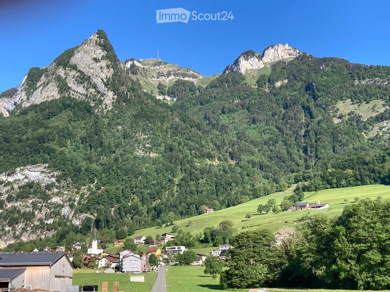 A picturesque village nestled in a lush green valley surrounded by towering mountains. The scene includes scattered houses, a church, and a clear blue sky.