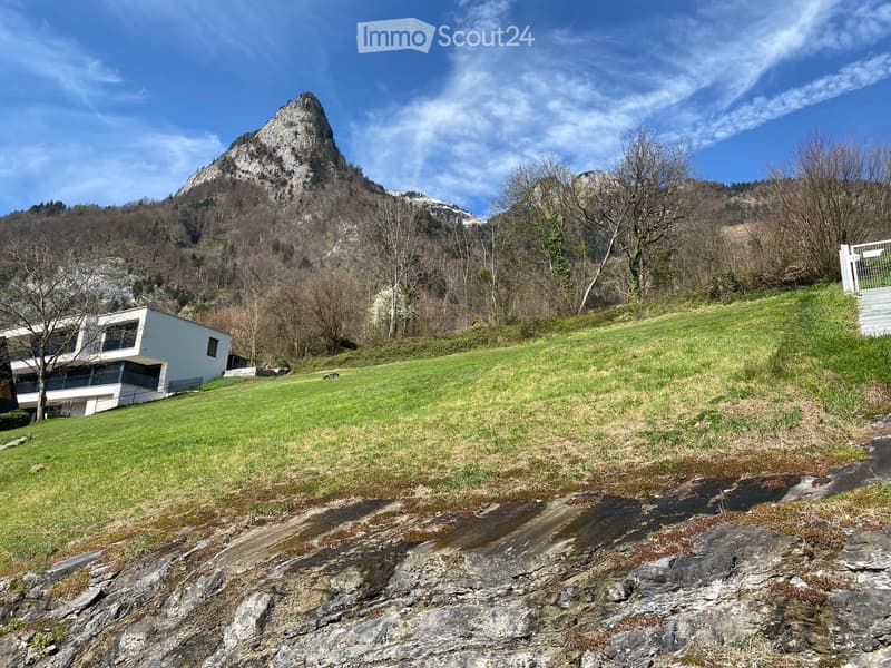 Modern white house with large windows and a balcony, situated on a grassy slope with a mountain backdrop.