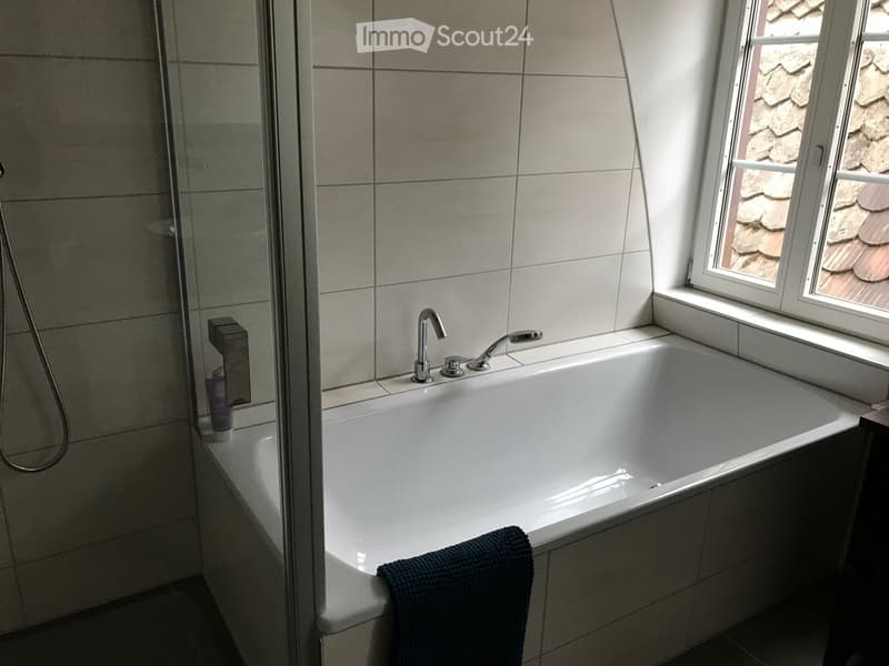 A bathtub with built-in shower head, faucet, and towel. Tiled walls, glass shower door, and a window. Shower gel and body lotion on the side.