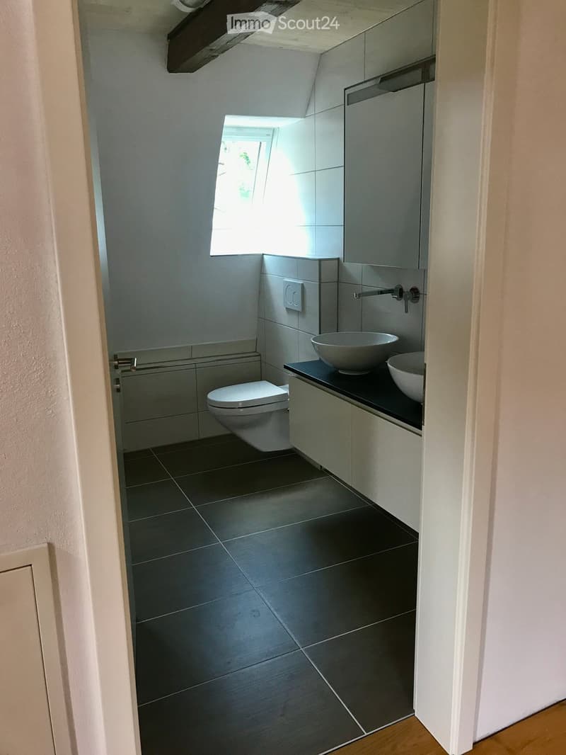 Bathroom with white toilet, double sink, mirror, tiled floor, small window, wooden ceiling beam