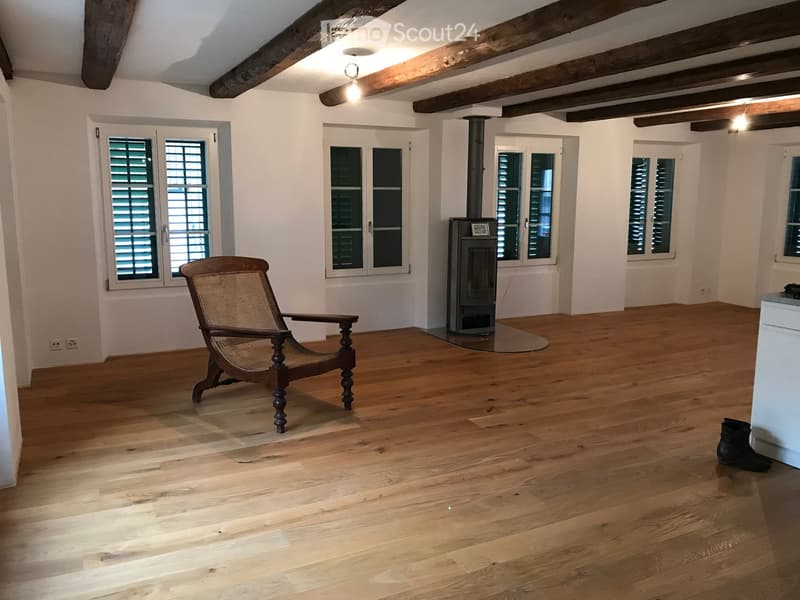 Empty room with wooden floor, white walls, 2 windows with wooden shutters, fireplace in the middle of the room, wooden chair, power outlets on the wall