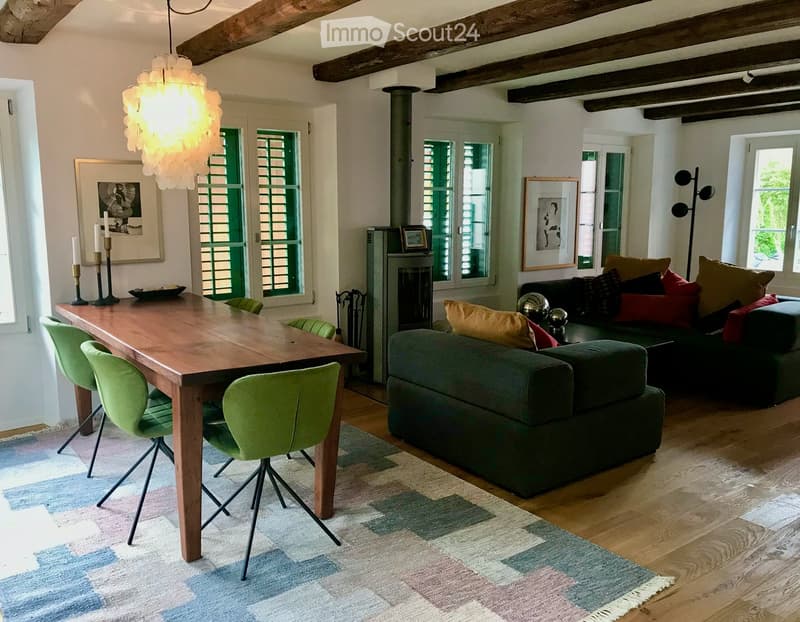 Dining room: large dining table with 4 green chairs, chandelier, patterned rug, fireplace, couch, 2 picture frames, lamp