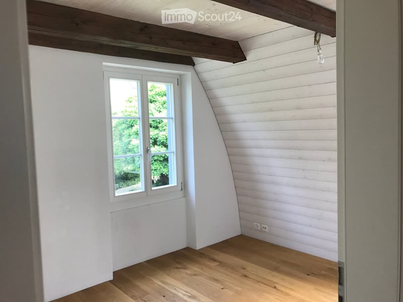 A narrow attic room with white walls, wooden floor, a window overlooking greenery, and wooden beams on the ceiling.
