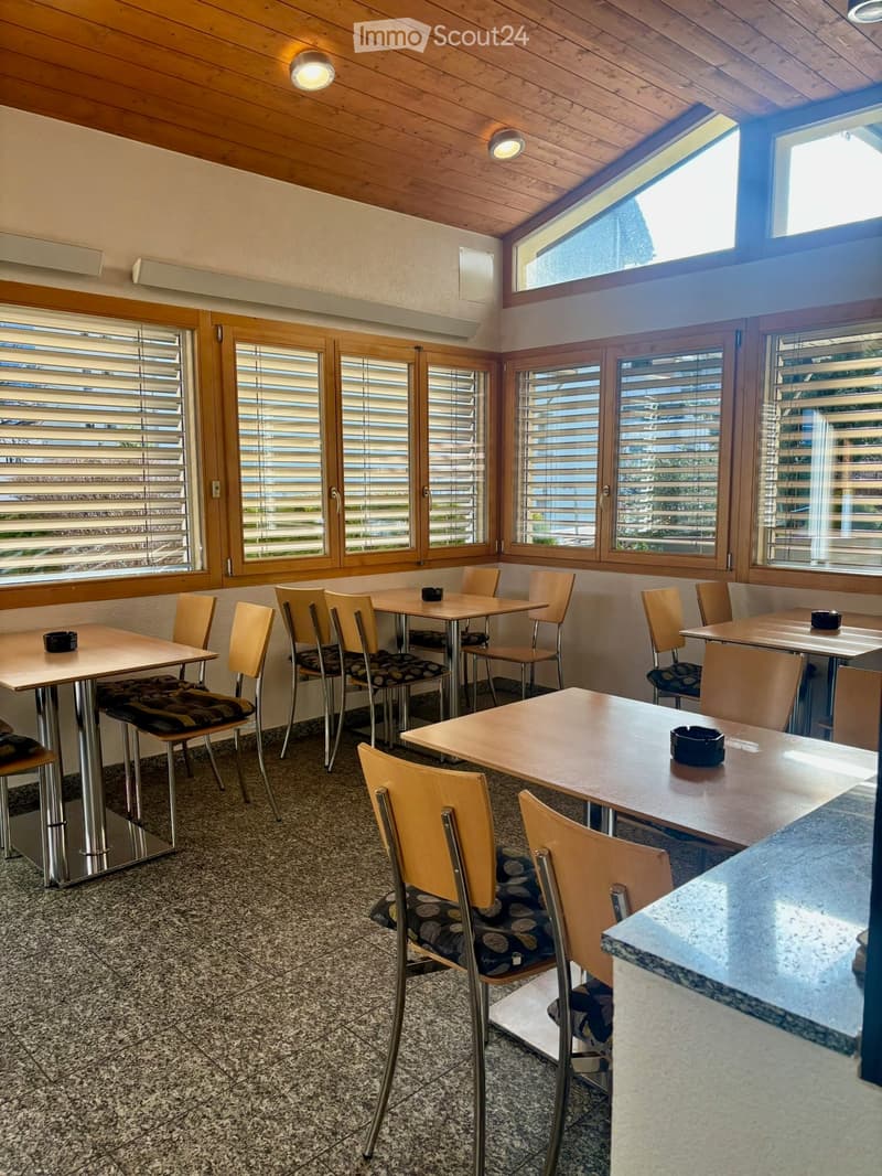Brightly lit dining area with wooden tables and chairs, large windows with blinds, and a polished tiled floor.
