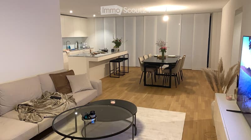 Modern open-plan living and dining area with a white sofa, glass coffee table, dining table with chairs, kitchen island, and television. The space features light wooden flooring and white walls.