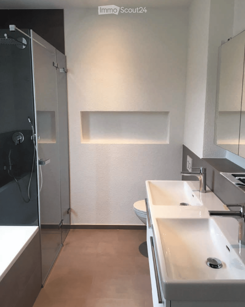 Modern bathroom with a glass-enclosed shower, two sinks, and a toilet. The walls are white with built-in lighting, and the floor is tiled.