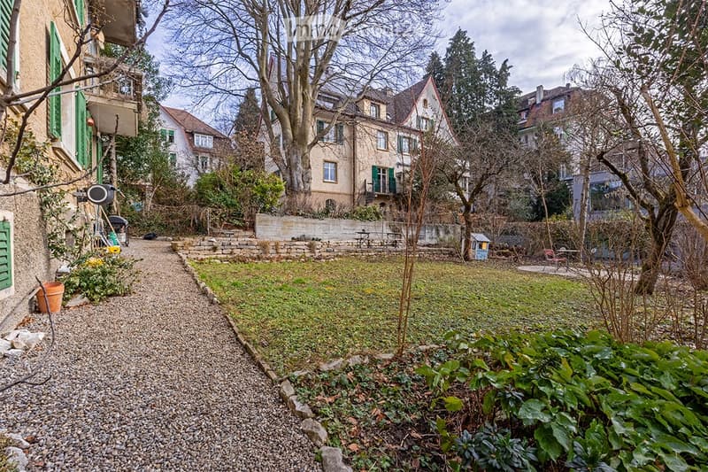 A small, well-maintained garden area with a gravel pathway, surrounded by trees and neighboring houses with green shutters. Features include a small shed, potted plants, and a stone wall boundary.