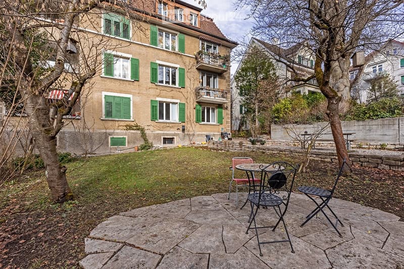 Garden area with a stone patio, outdoor furniture, and a multi-story building with green shutters in the background.