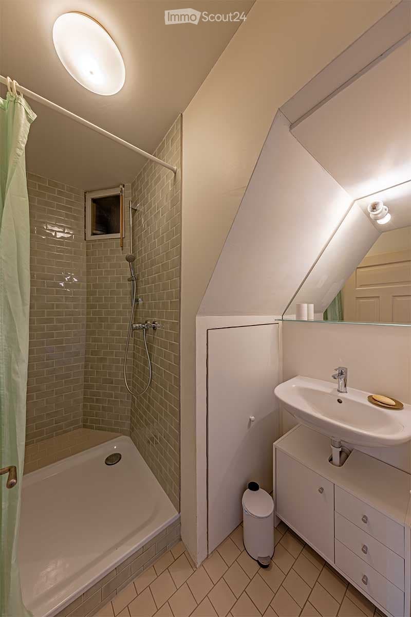 The bathroom has a shower with glass doors, a bathtub, a sink with a faucet, a mirror, white tiles, and a waste bin. It also has a small window for ventilation.