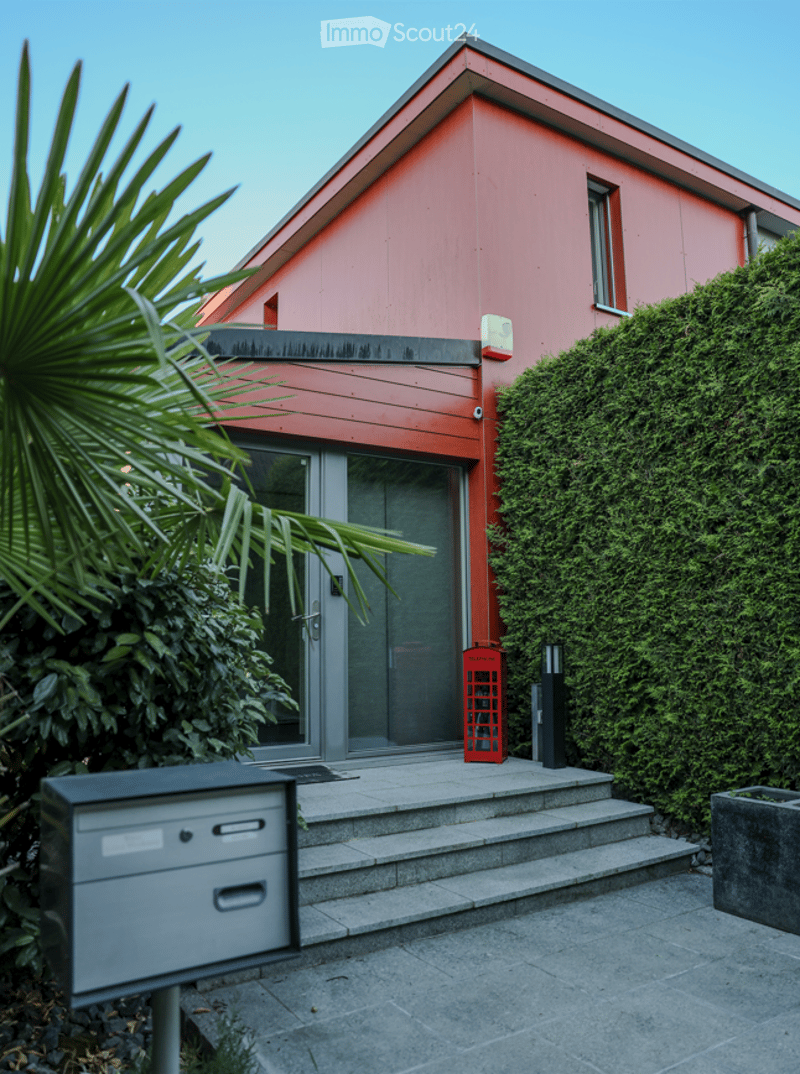 Modern two-story house with red exterior, large glass front door, small porch with steps, and surrounding greenery.