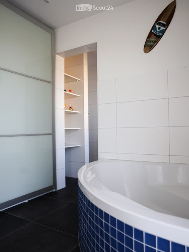 Modern bathroom with a white bathtub featuring blue tiles, dark tiled floor, and an open shelf unit with various items.