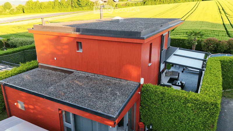 Modern two-story house with red exterior and grey gravel roofs, featuring a patio with outdoor furniture and a swimming pool.