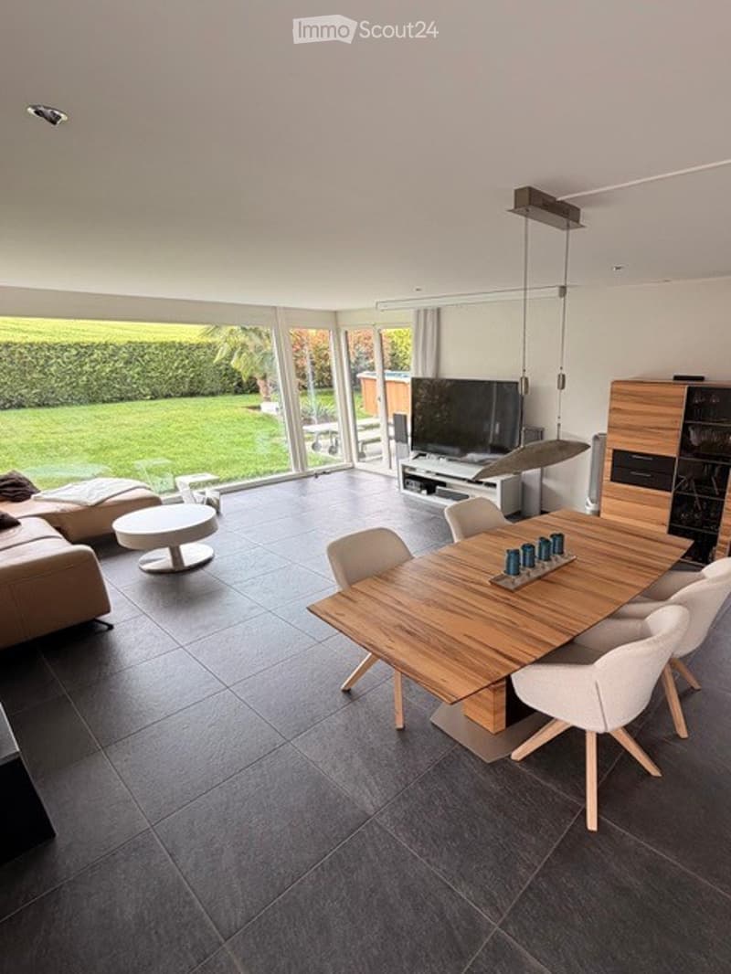 Open-plan living and dining area with large windows offering a view of a green garden. Features a grey sofa, coffee table, wooden dining table with white chairs, and a TV on a stand.