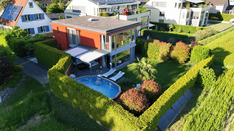Aerial view of a modern house with a swimming pool, surrounded by well-maintained hedges and greenery, with neighboring houses visible in the background.