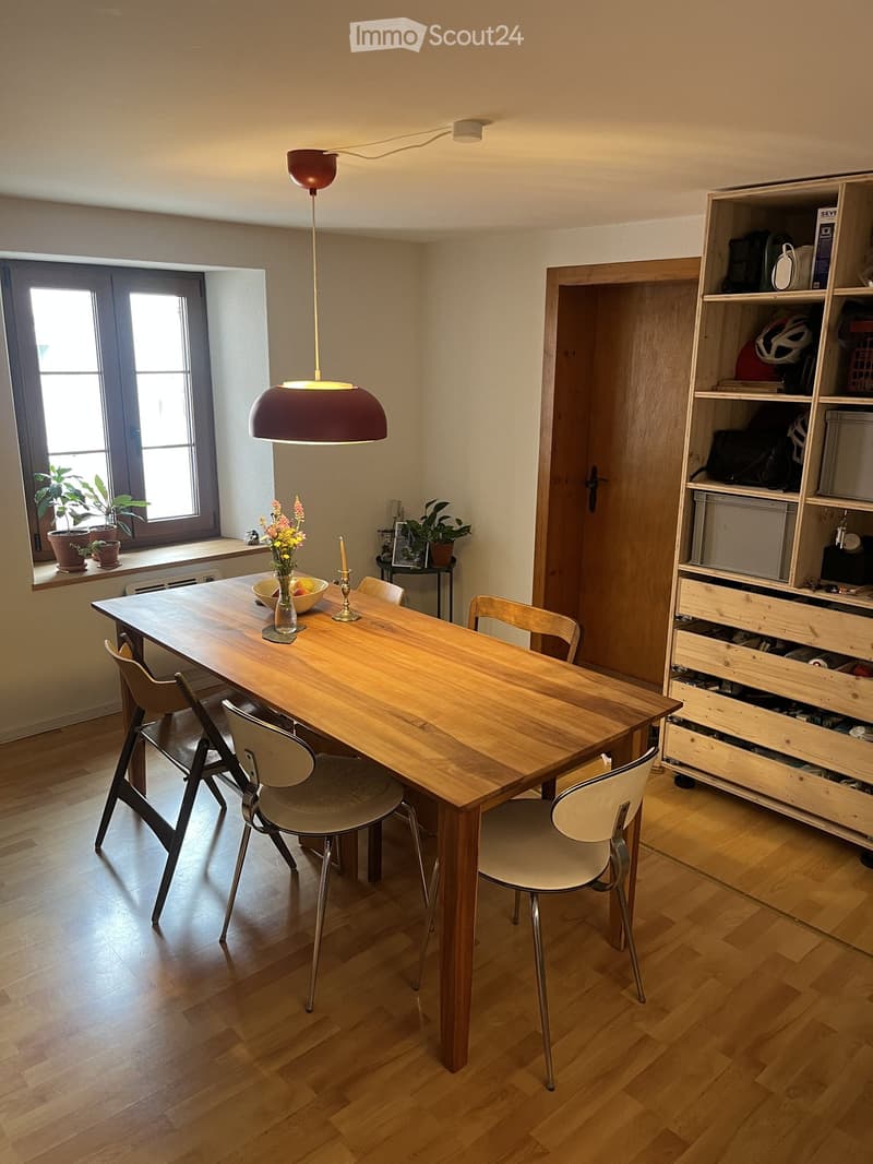 Dining area with a wooden table, six chairs, a vase with flowers, a bowl of fruit, a candle, and a wooden shelf with storage compartments and various items.