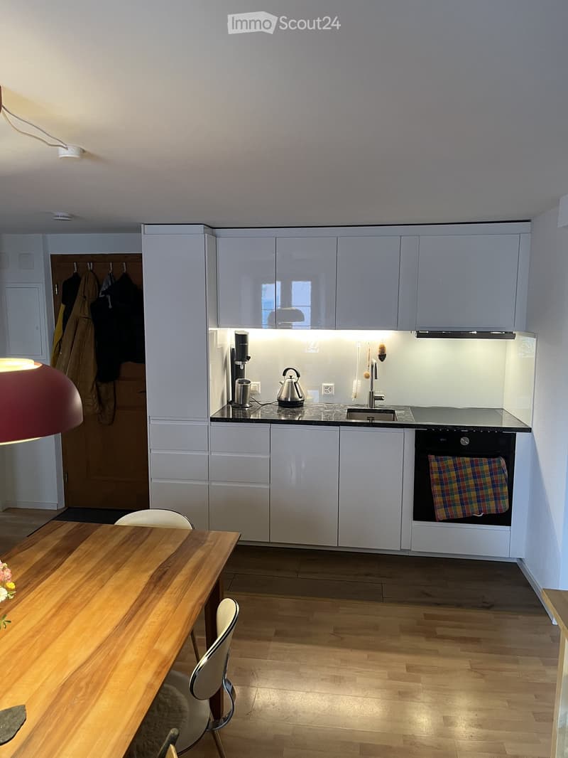 Modern kitchen with white cabinets, black countertop, sink, oven, coffee maker, teapot, and a wooden dining table with chairs.