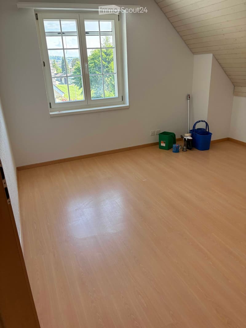 Empty room with wooden flooring, white walls, and a large window showing a view of greenery outside. Cleaning supplies are placed near the window.