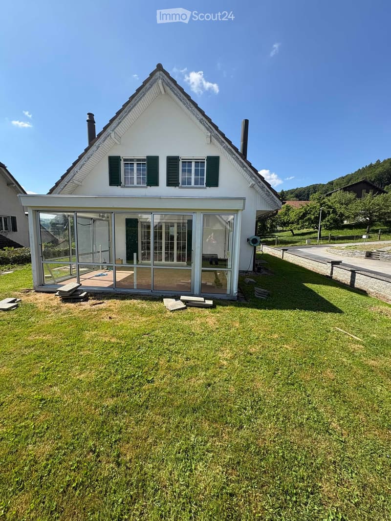 Two-story white house with green shutters, large glass sliding doors leading to a patio, surrounded by a well-maintained lawn and trees.