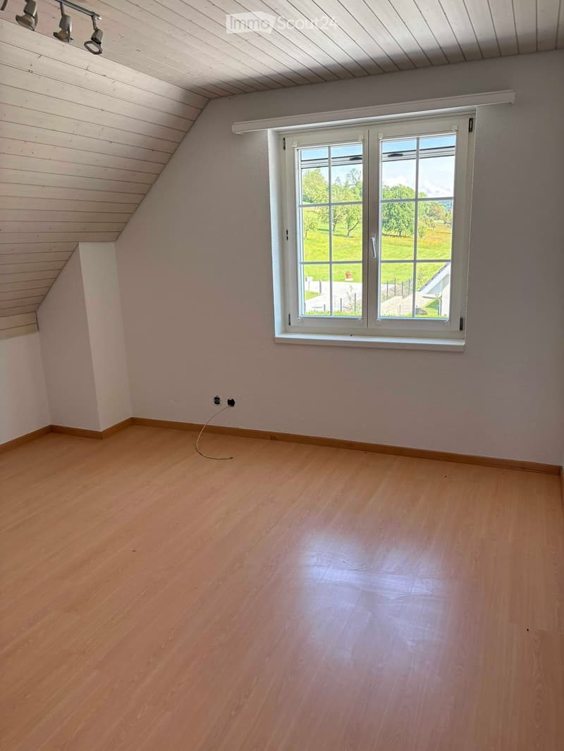 Empty room with wooden flooring, white walls, and a large window offering a view of a green landscape.