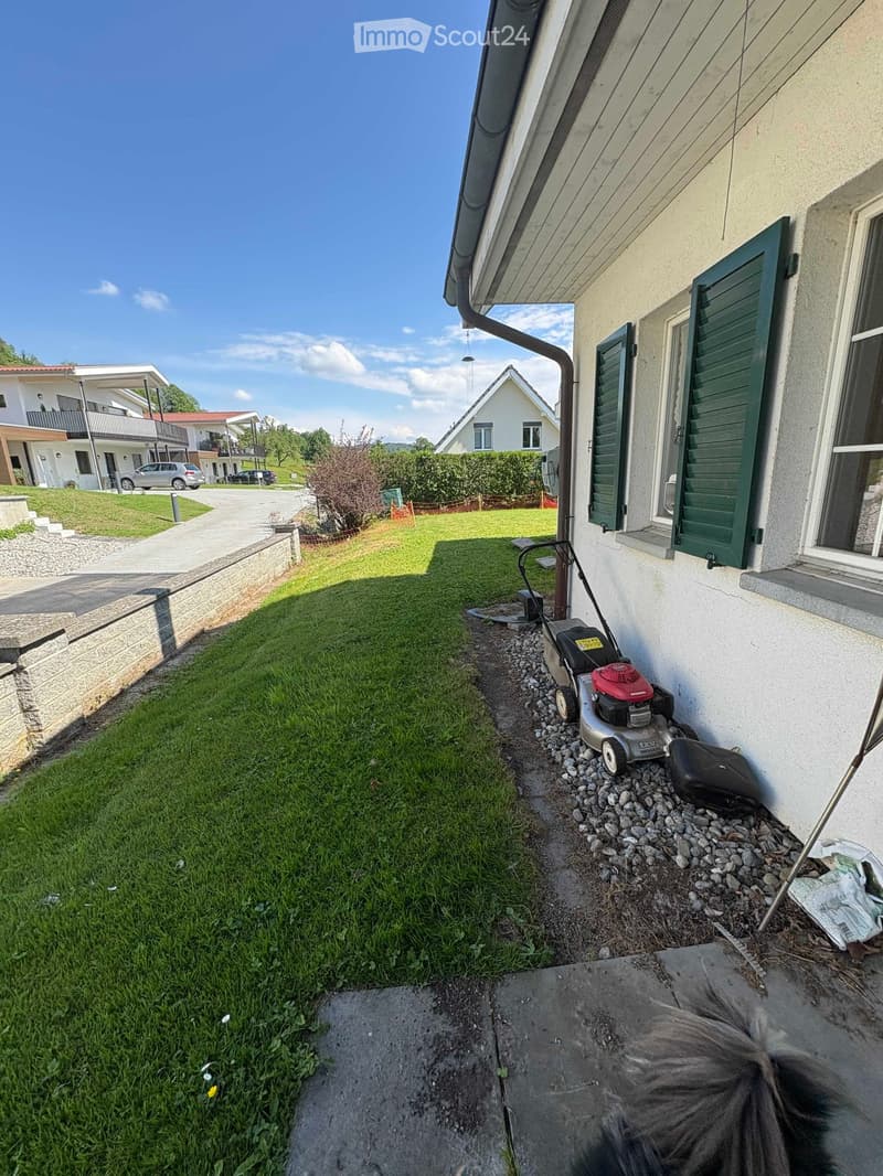 Lawn mower placed on a gravel path next to a house with green shutters. The surrounding area includes a well-maintained lawn and a paved driveway leading to neighboring houses.