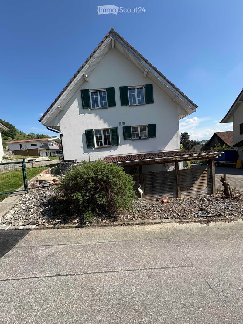 Two-story house with white exterior, green shutters, and a pitched roof. The house features multiple windows and a small covered entrance area with a wooden fence and decorative rocks.